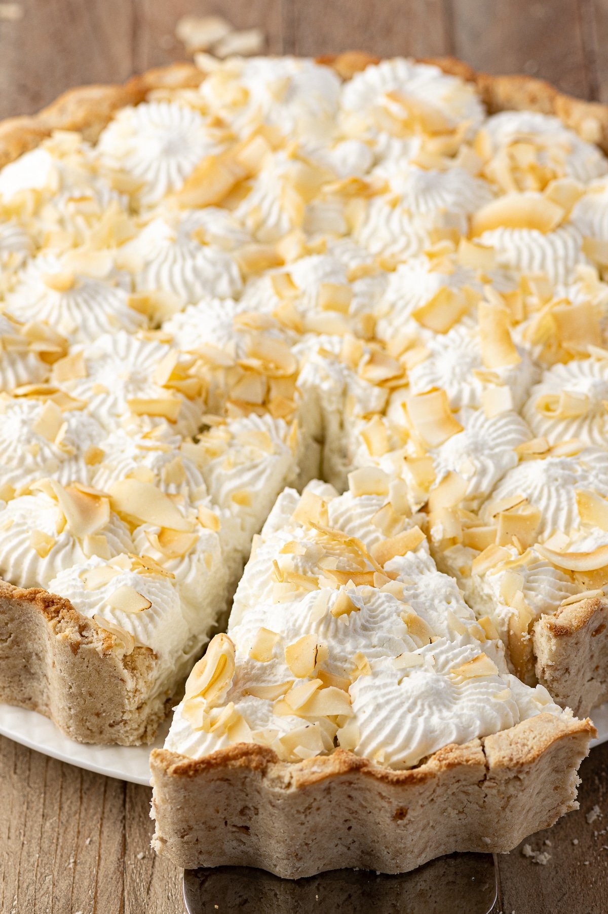 Coconut cream pie with a slice being removed on a rustic wooden table. 