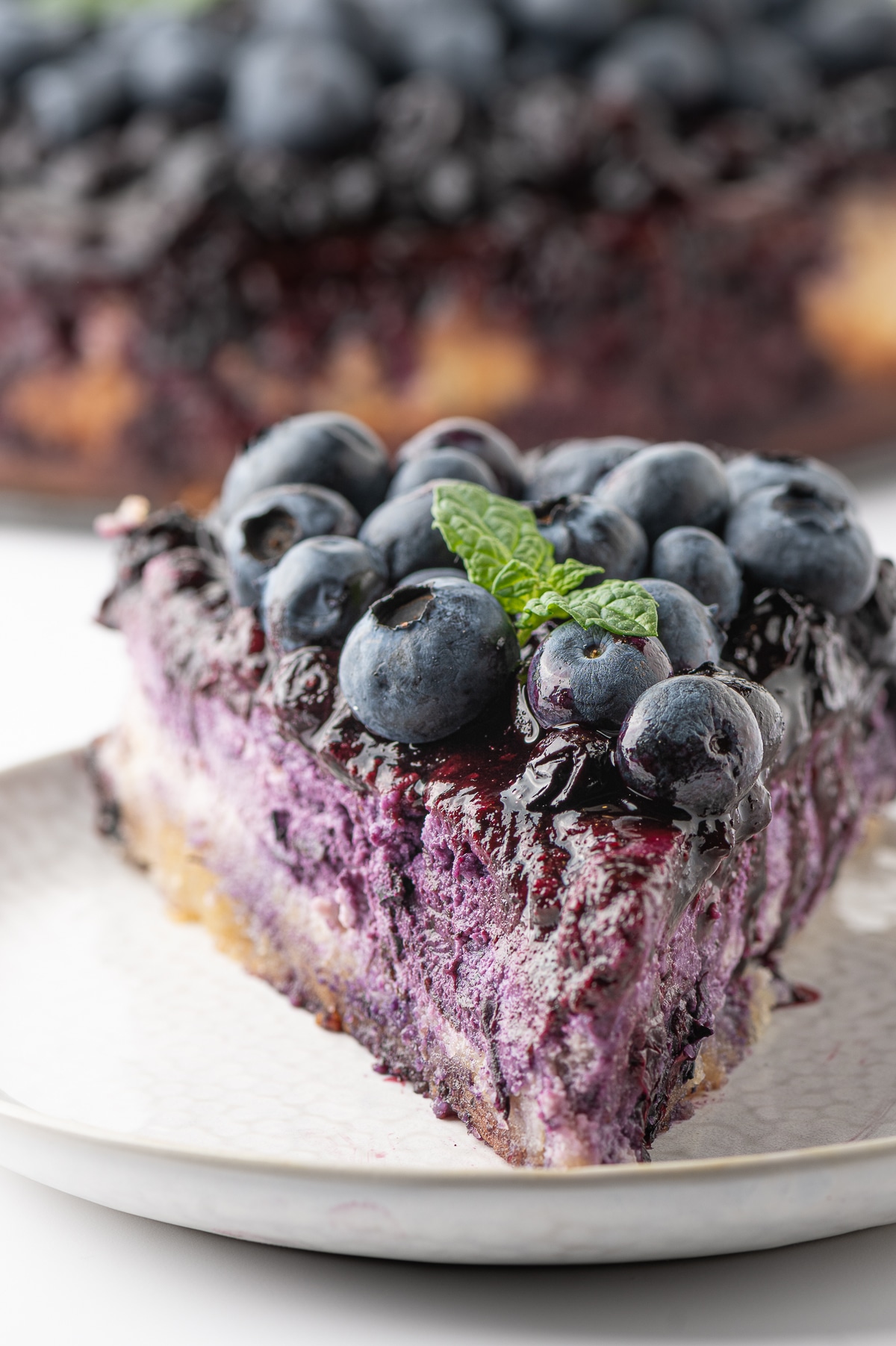 An up close photo of a piece of bright violet colored cheesecake topped with blueberry drizzle, fresh big bright blueberries and a sprig of bright green mint