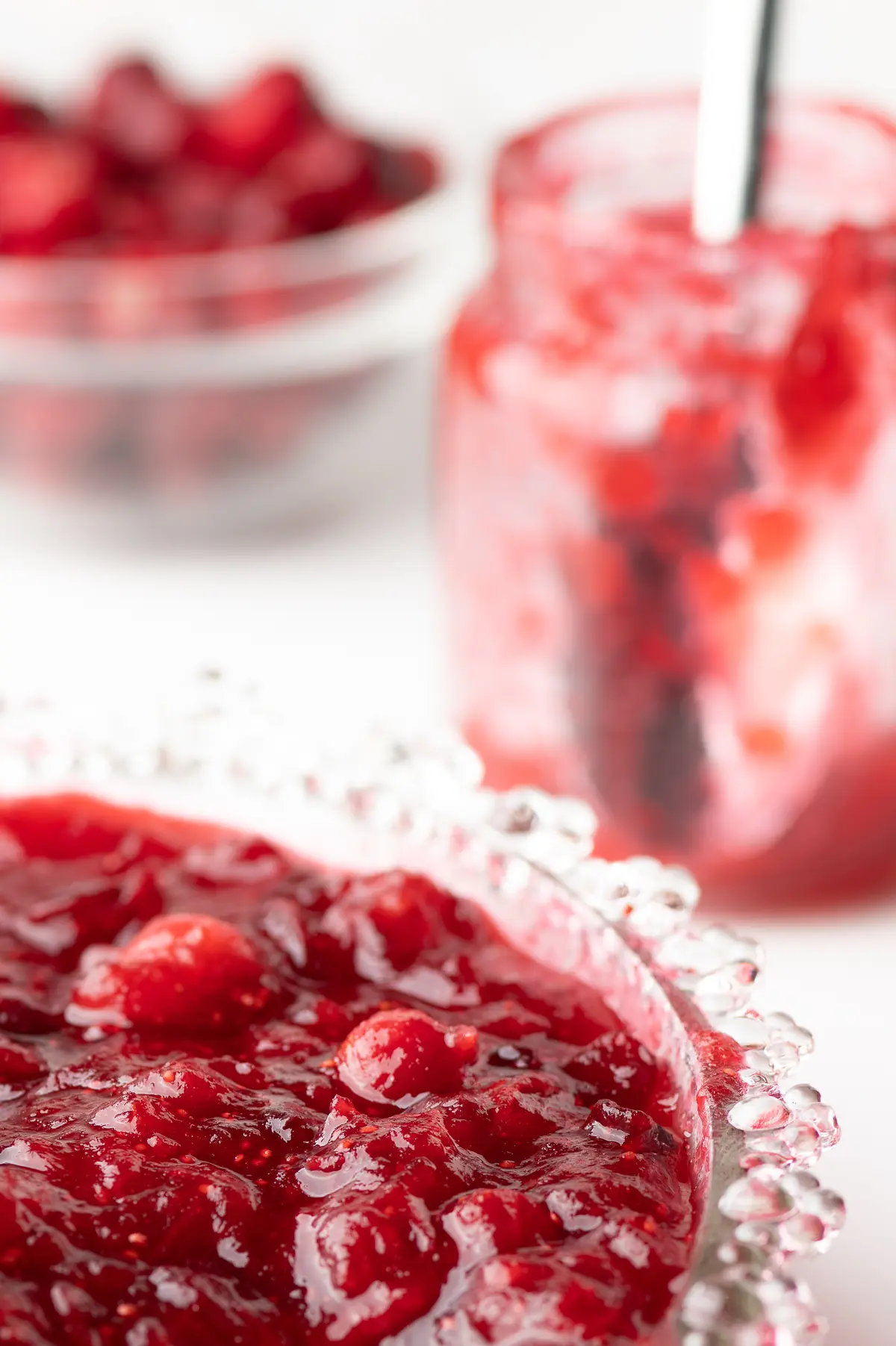 A bowl filled with bright red cranberry sauce and an empty jar with a spoon all against a bright white background