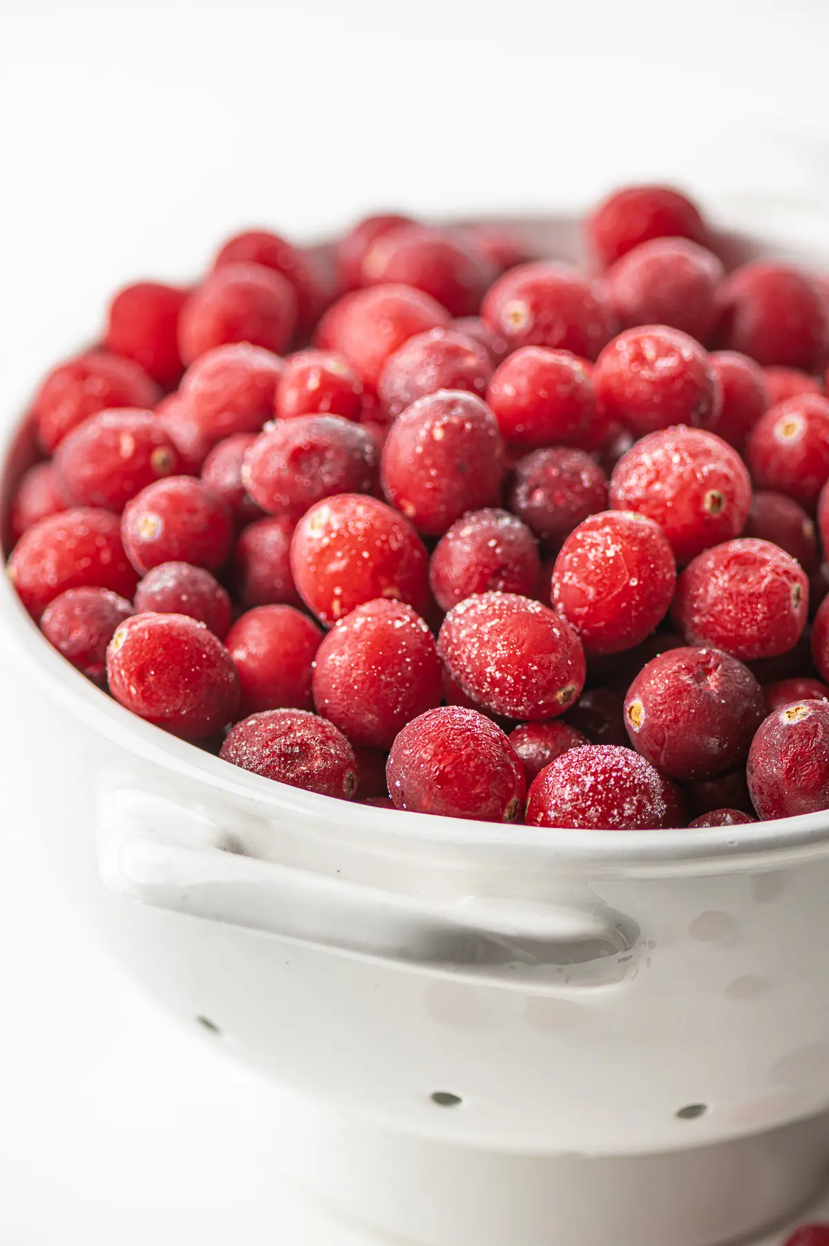 A small white bowl filled with icy frozen cranberries all on a bright white background .