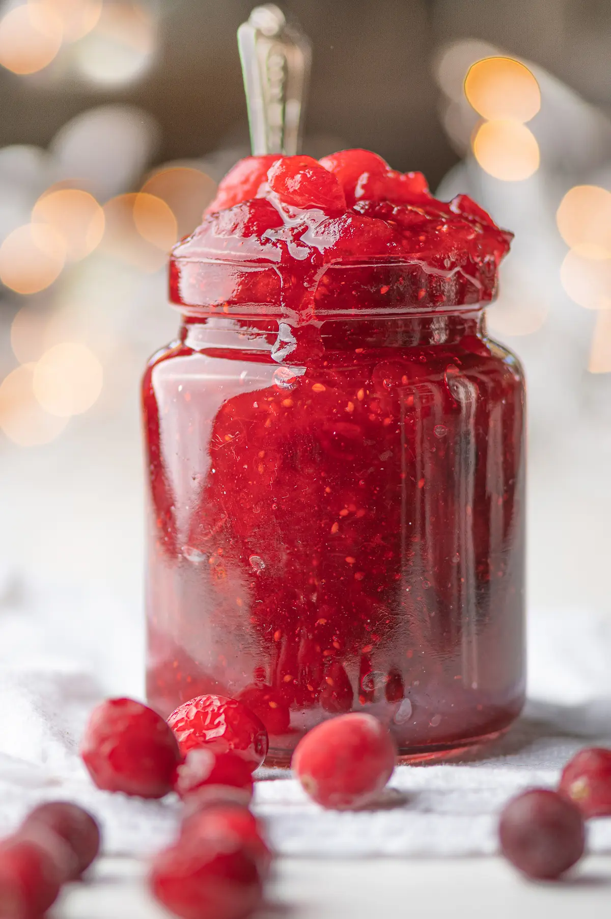 A jar filled with bright red cranberry jelly with scattered cranberries around the base of the jar.  In the background there are blurred out Christmas lights