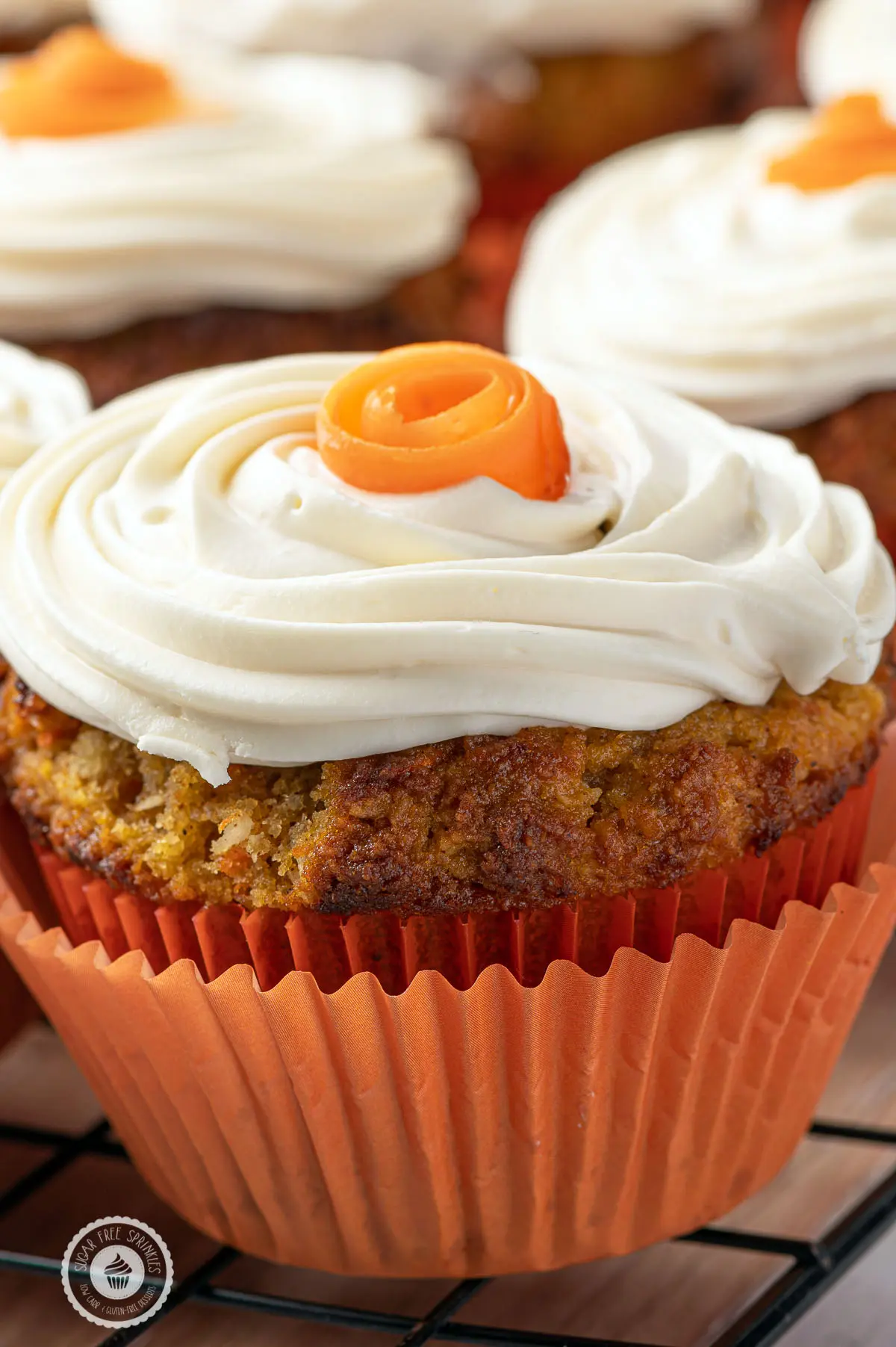 Close up of a low carb carrot cake muffins in an orange muffin rapper with swirls of cream cheese frosting and small orange roses on the top. 