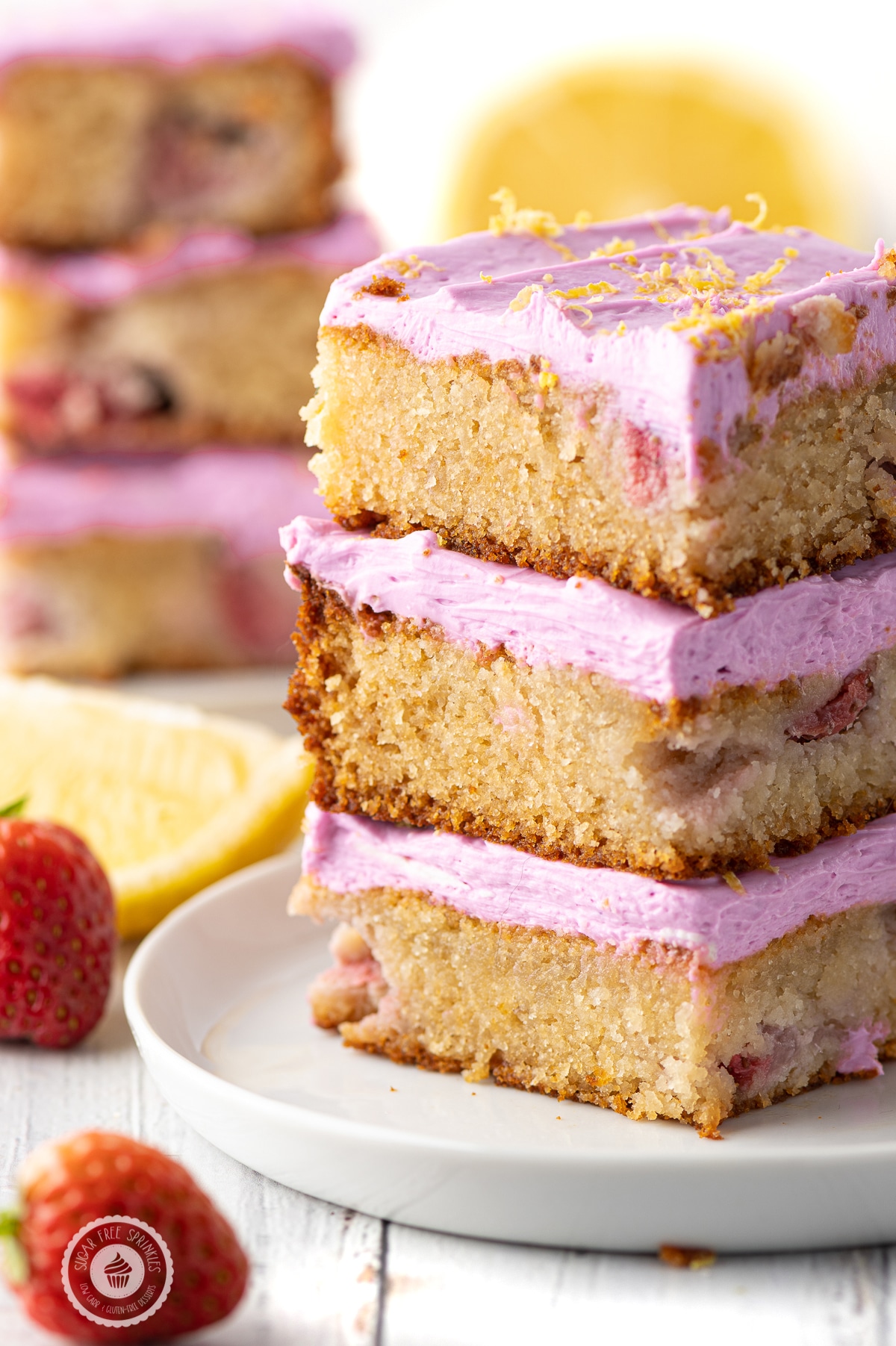Three stacked low carb strawberry lemon blondies with pink frosting on a white plate with lemon wedges  and strawberries .