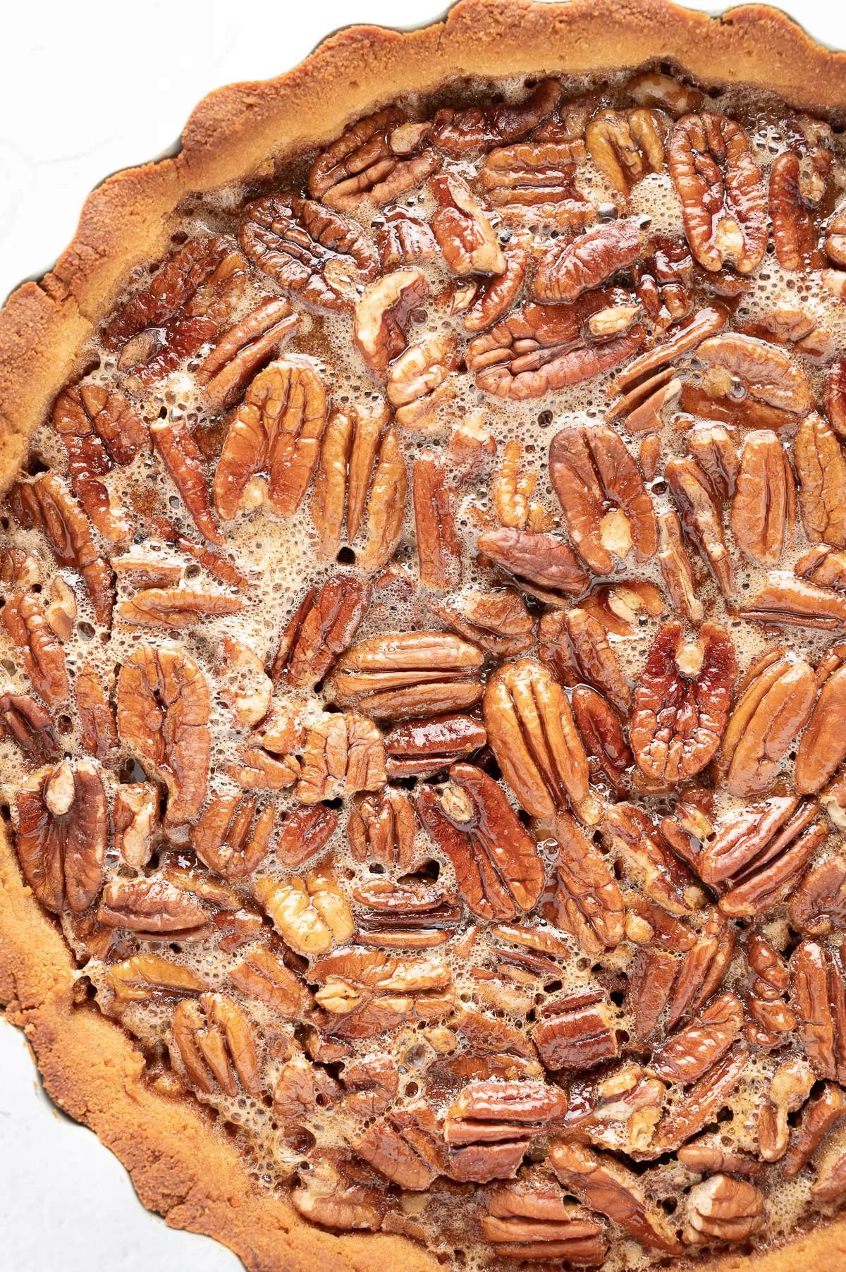 a close up image of the top of a pecan pie showing the sticky gooey texture of the filling and golden baked pecans in a perfectly baked crust. 