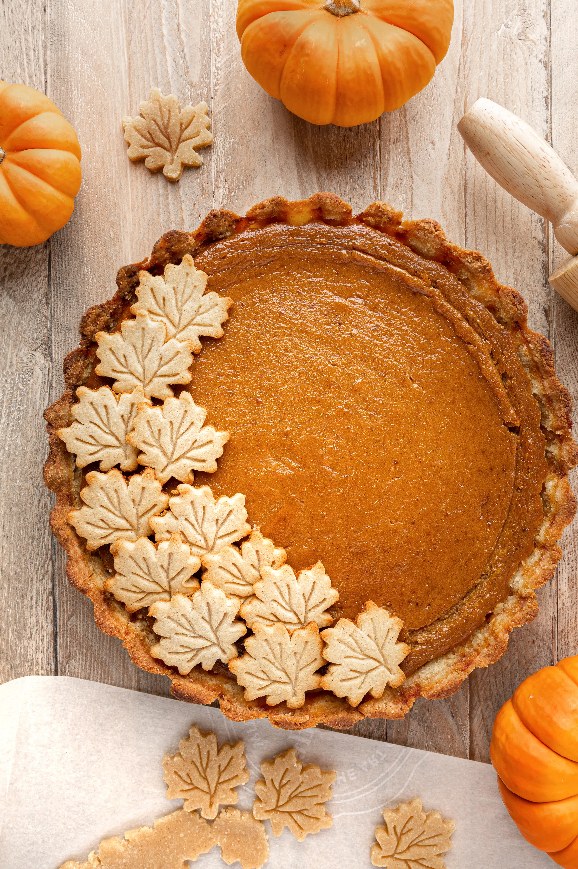 Pumpkin pie with maple shaped decorations on a rustic wooden background surrounded by mini orange pumpkins. 