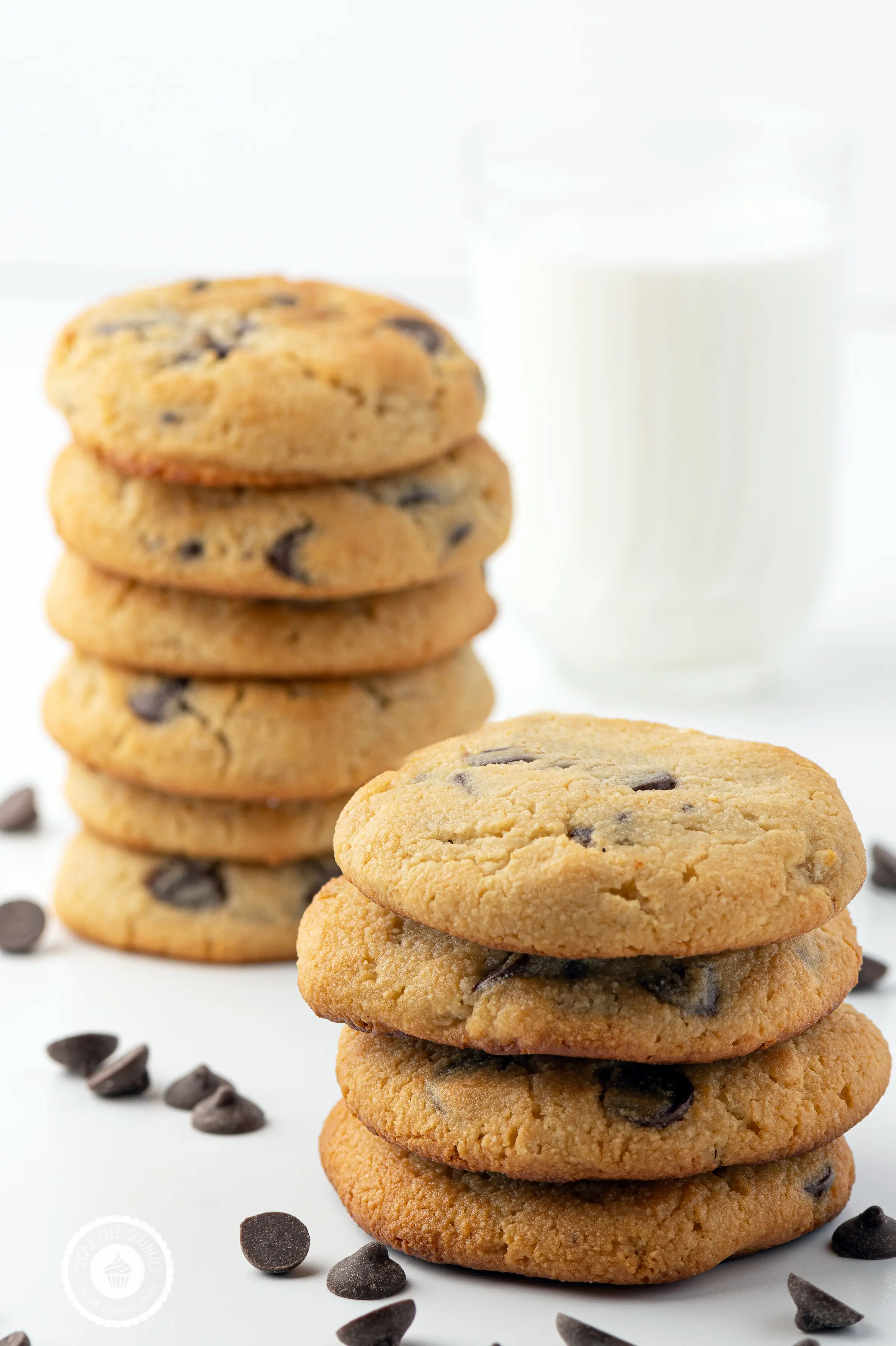 Stacks of keto chocolate chip cookies on a white background with scattered chocolate chips and a glass in the background filled with milk. 