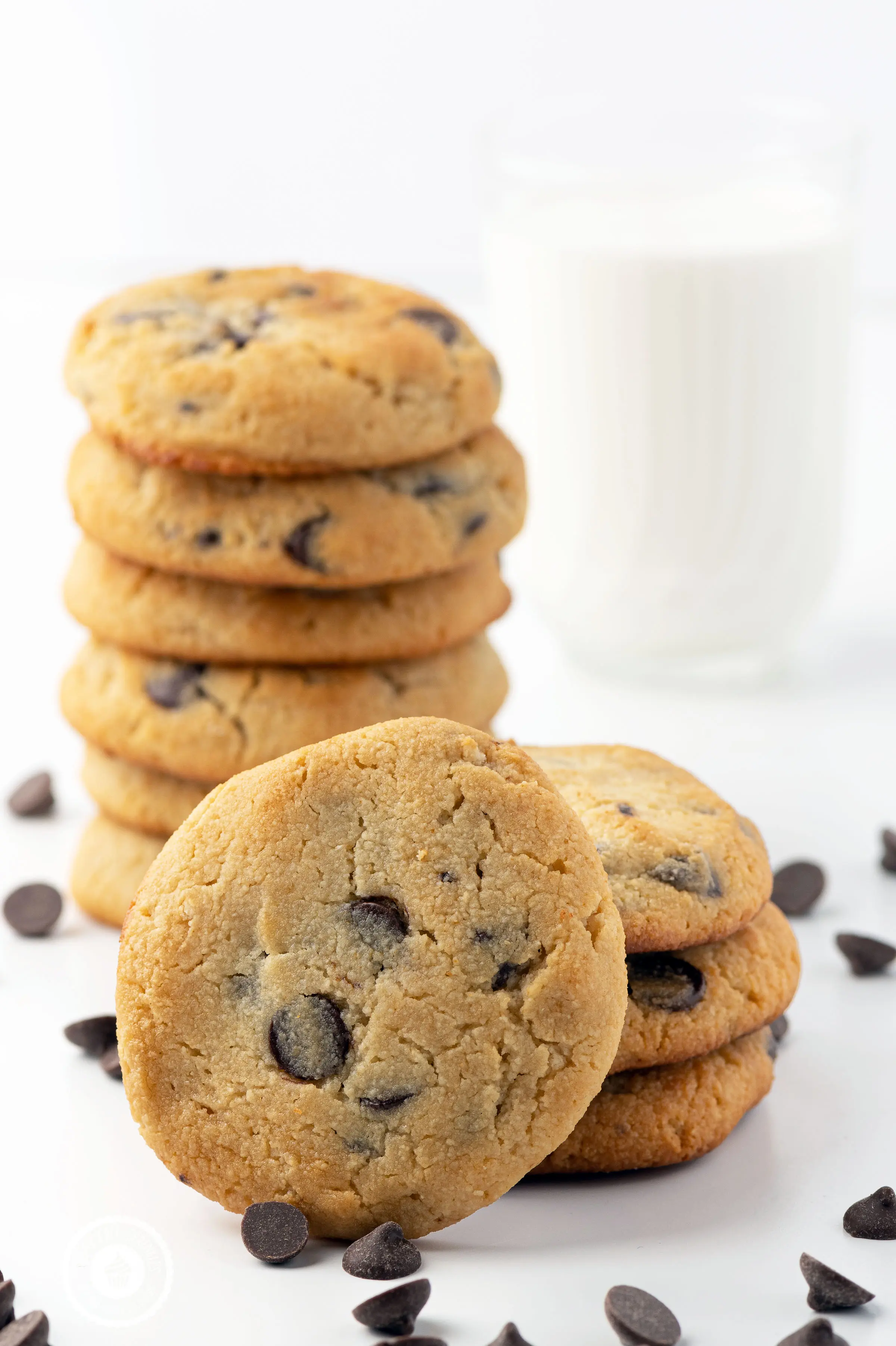 Stacks of keto chocolate chip cookies on a white background with scattered chocolate chips and a glass in the background filled with almond milk. 