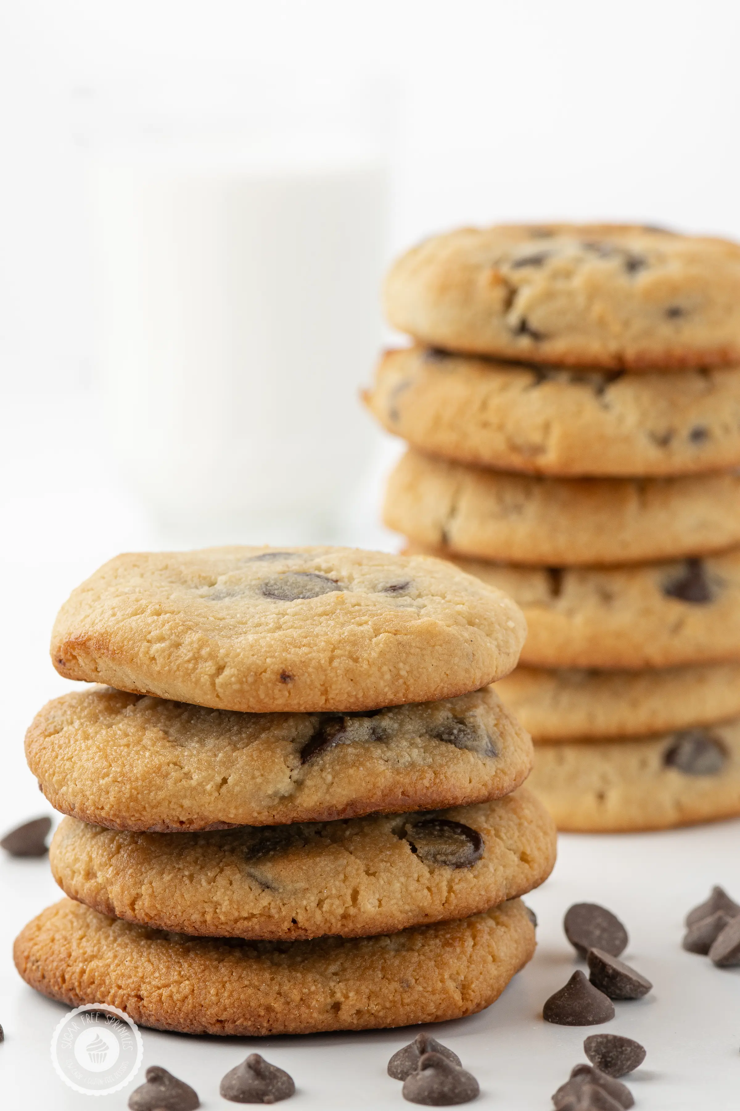 Stacks of keto chocolate chip cookies on a white background with scattered chocolate chips and a glass in the background filled with almond milk. 