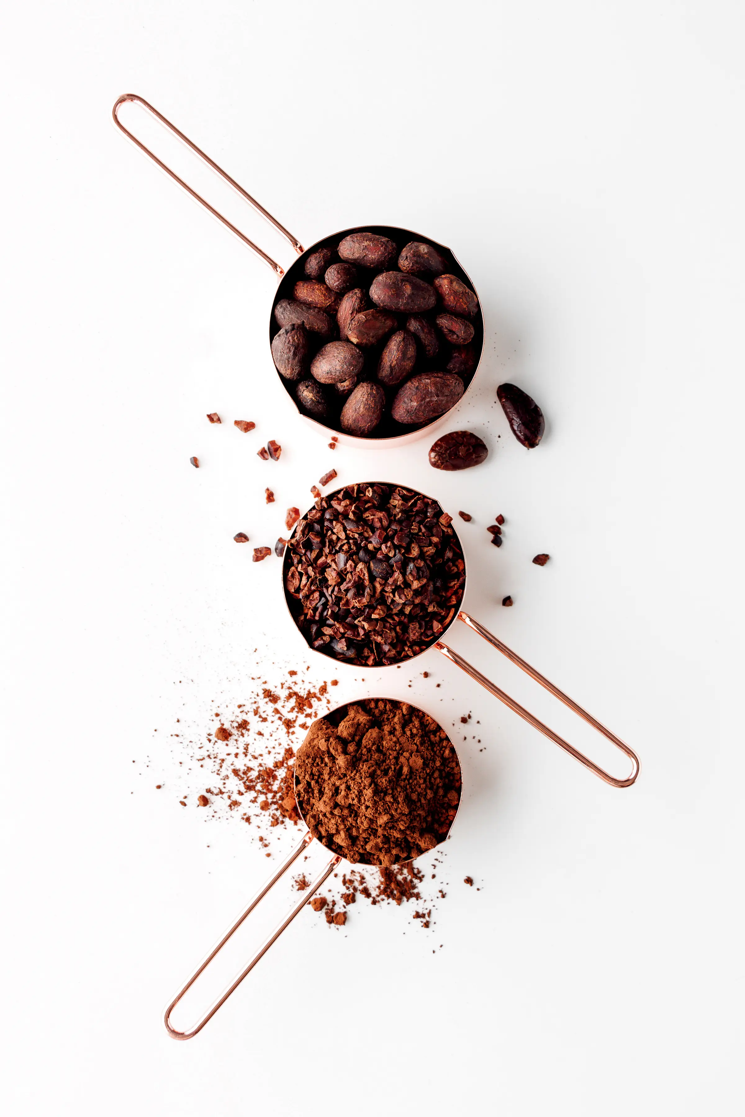 Three measuring cups with cocoa powder, cocoa nibs and cocoa beans on a bright white tabletop. 