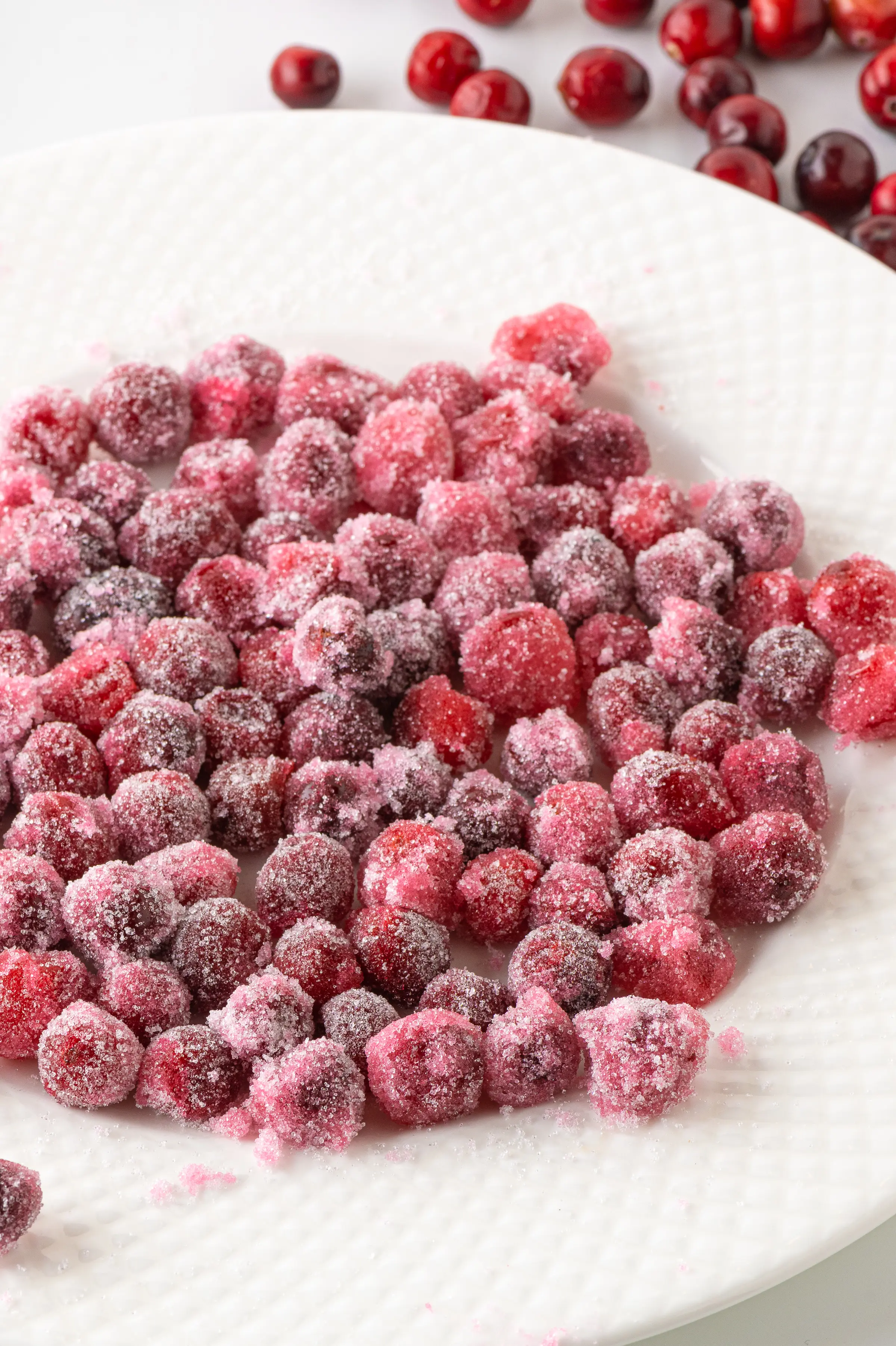 A white plate filled with red ripe cranberries coated in granular sweetener.  The berries look like they have been covered in a pink winter frost. 