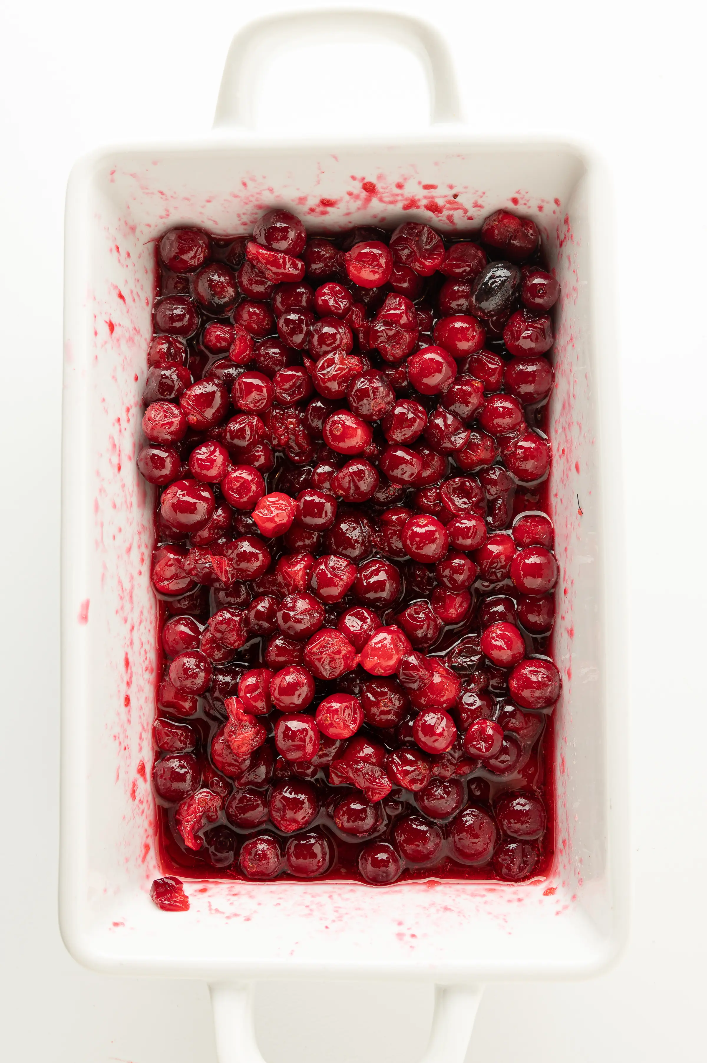 A white dish filled with ruby red berries glossy coated with a thick syrup. 