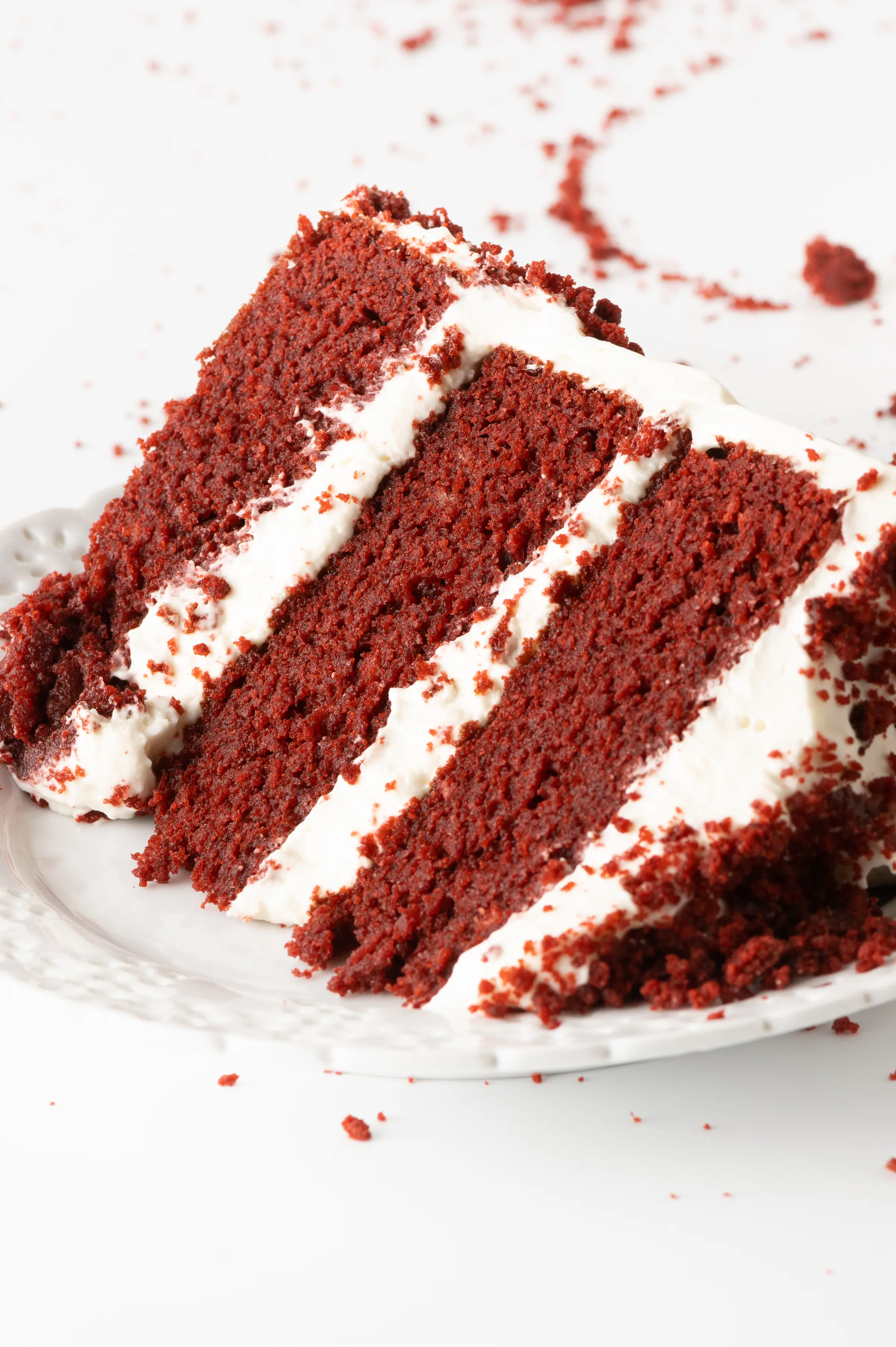 A single slice of bright red maroon red velvet cake with bright white layers of frosting.  The cake is set on a white texture plate and set against a bright white tabletop with red cake crumbs scattered around the plate. 