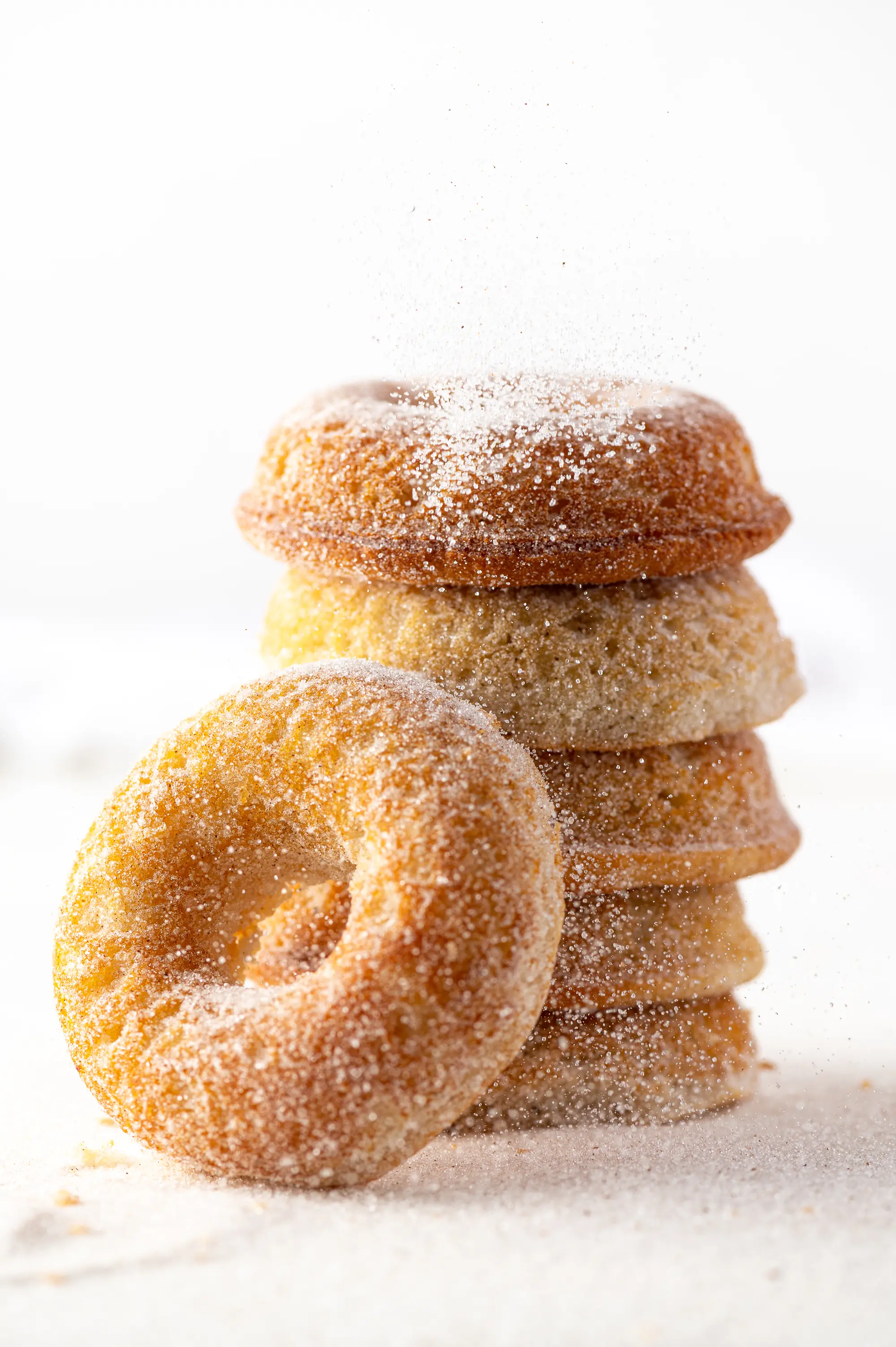Old fashioned gluten free donuts stacked against a bright white background with cinnamon sugar being sprinkled over the top. 