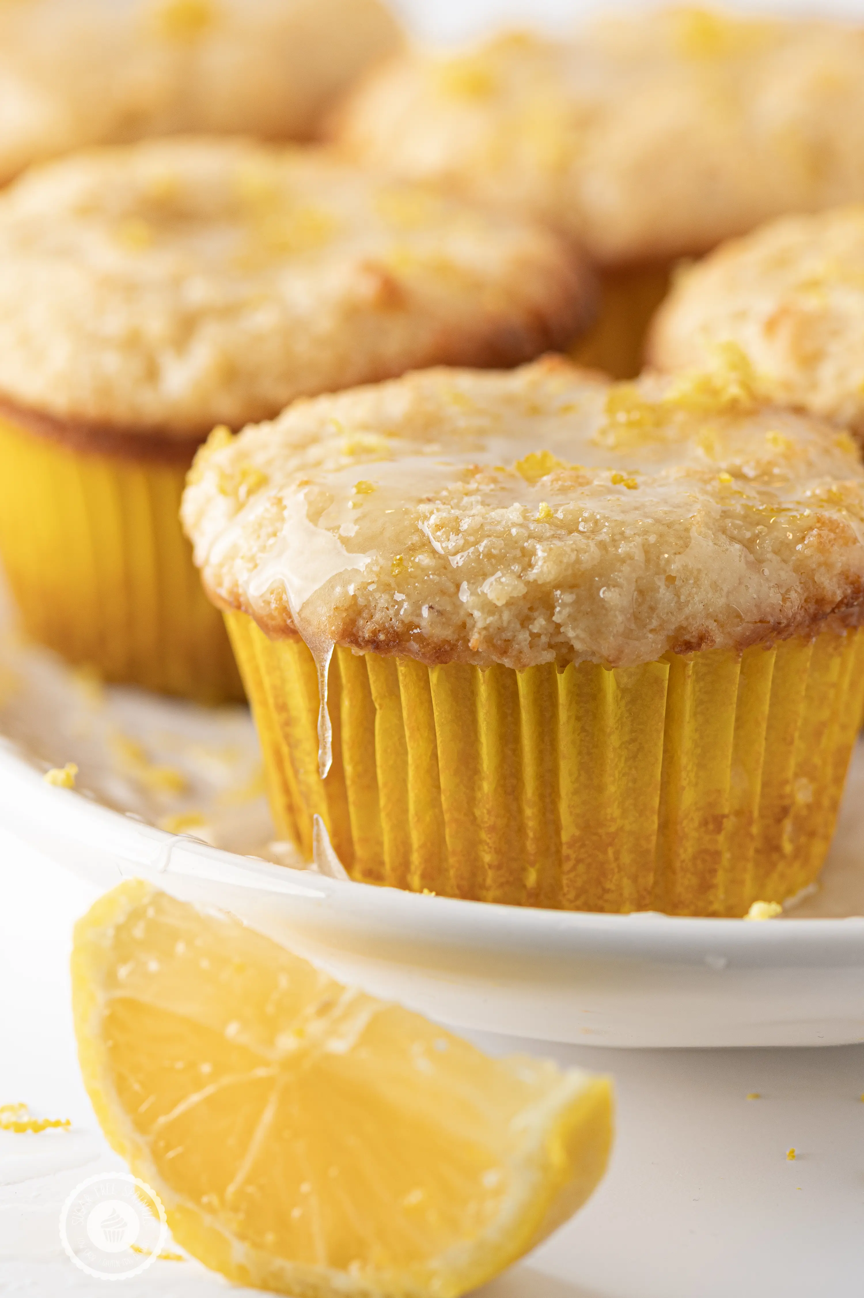 Lemon muffin in a yellow muffin liner, dripping with sticky glaze and topped with fresh lemon rind.  Muffins are stacked in behind slightly out of focus. 