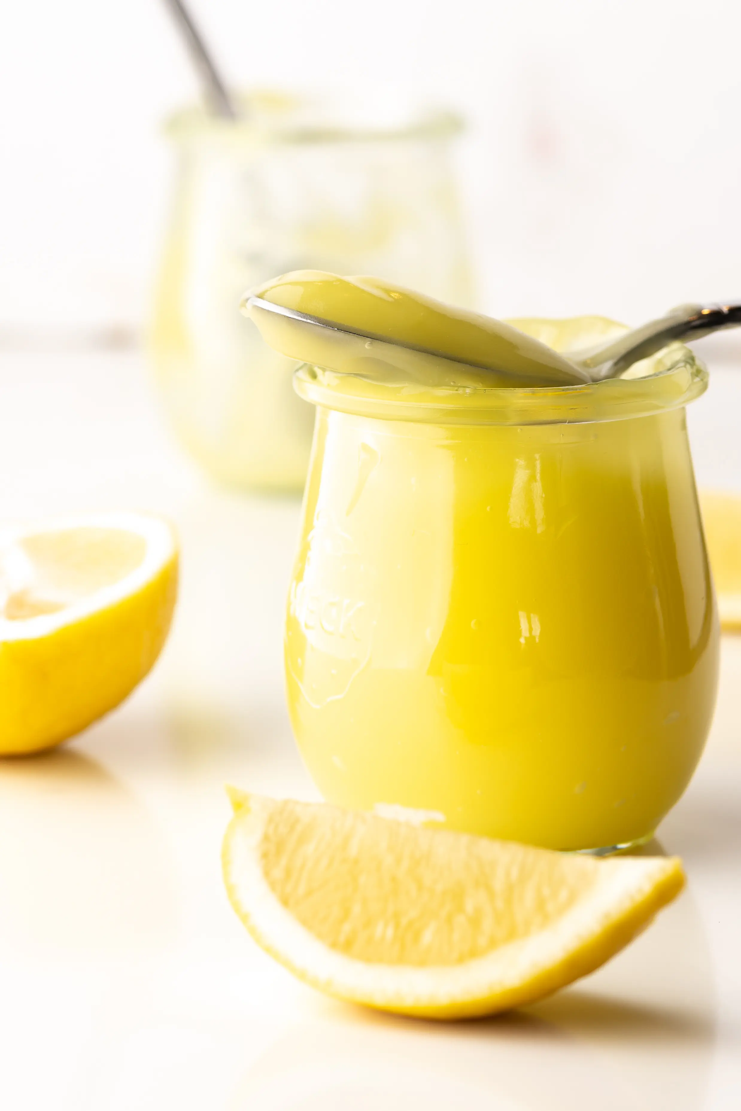 Bright yellow keto lemon curd in a small jar surrounded by pieces of sliced lemons.  An empty jar with a spoon is in the background. 