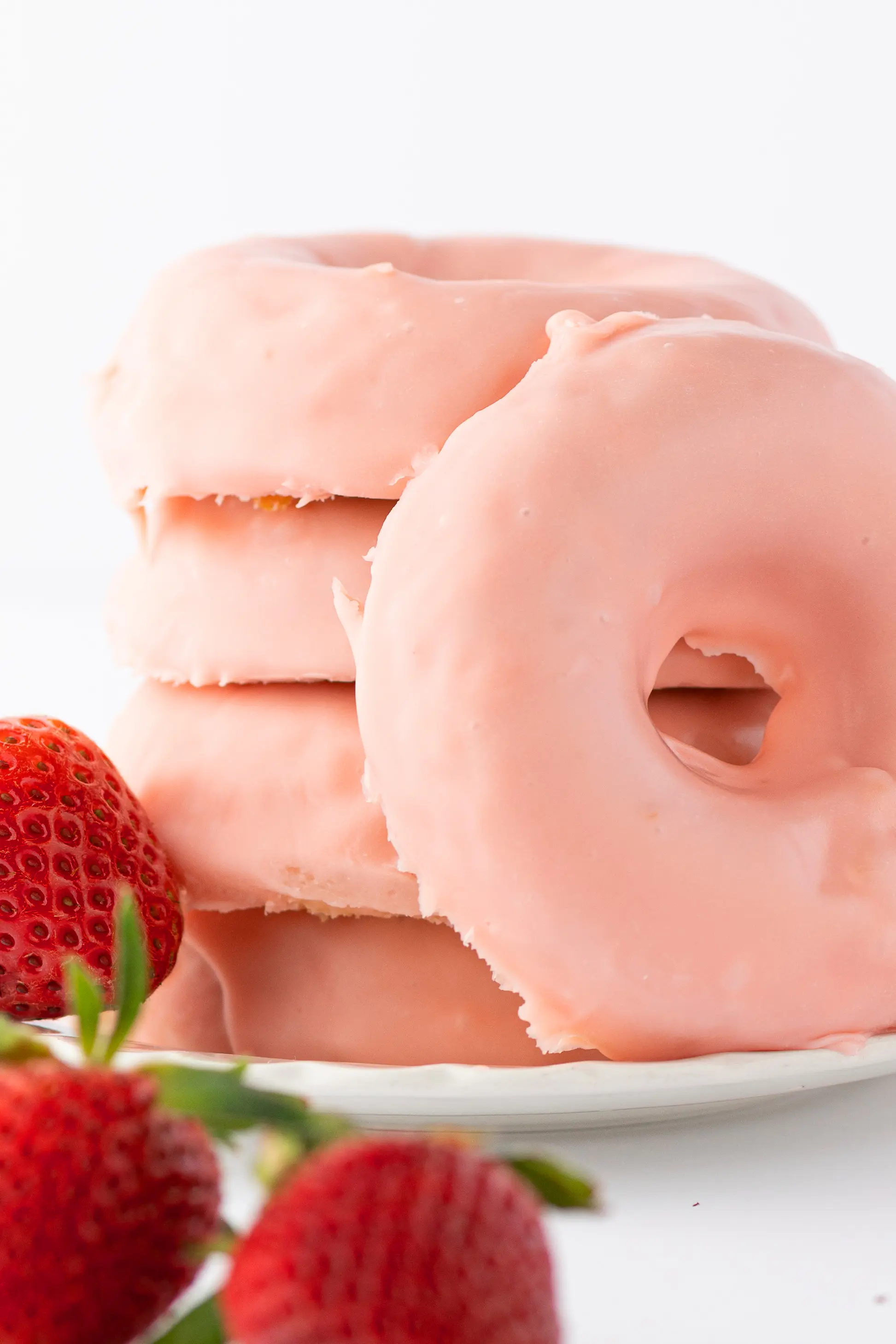 A stack of strawberry glazed donuts on a white plate with fresh ripe red berries scattered around the tabletop.