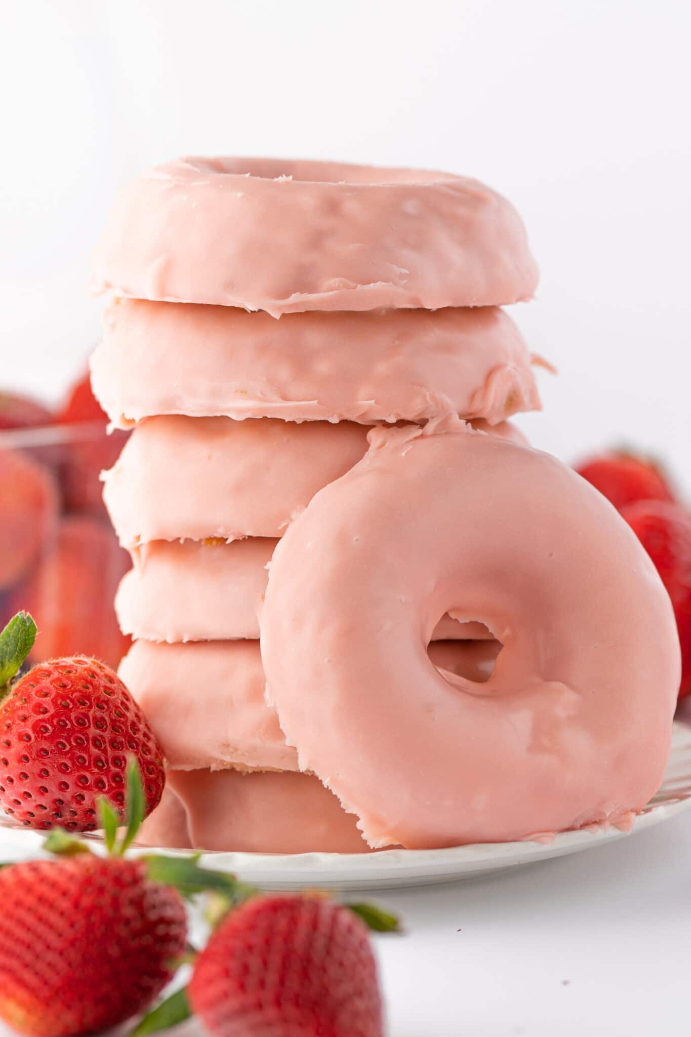 A stack of keto strawberry donuts on a white plate with fresh ripe red berries scattered around the tabletop.
