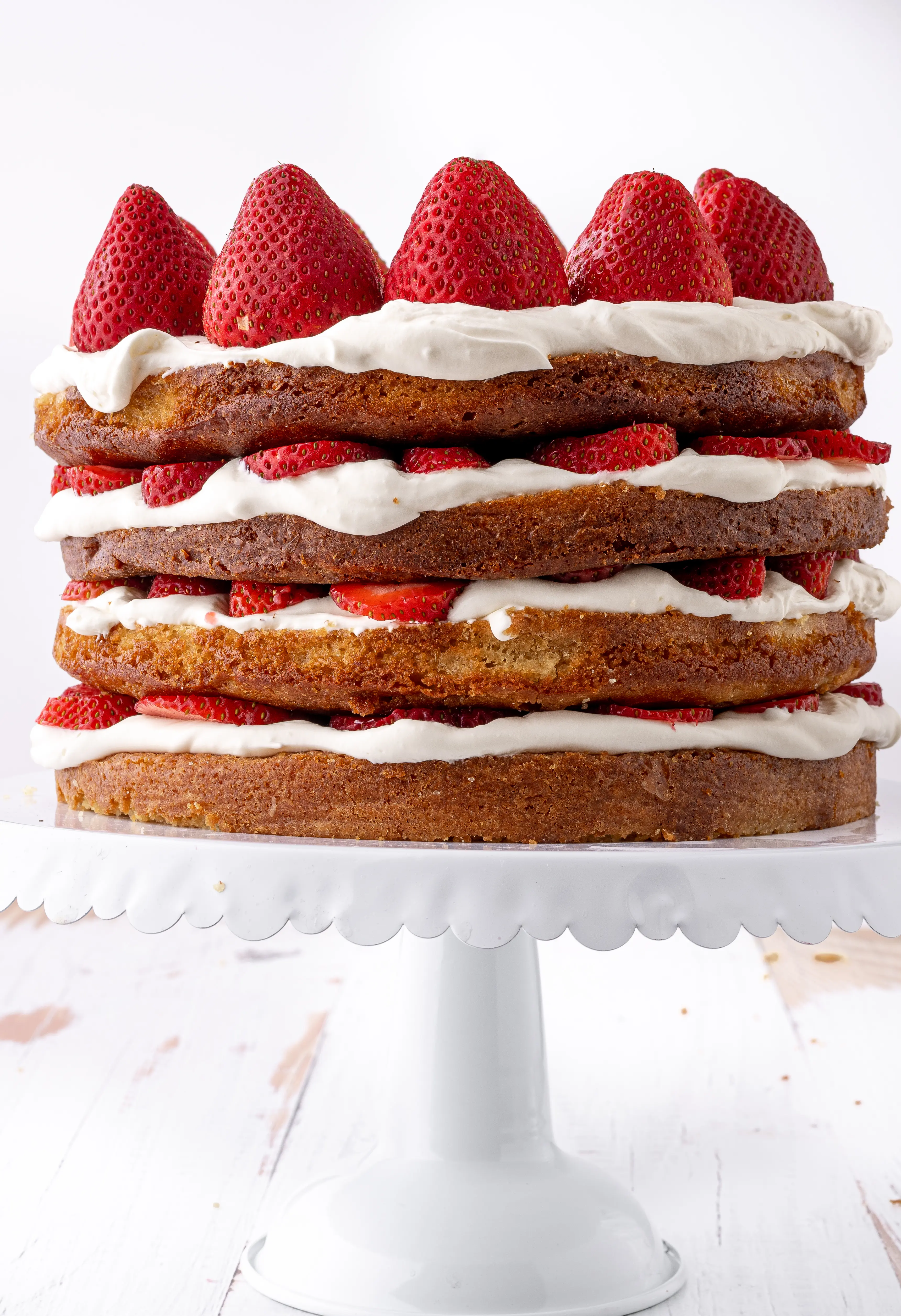 A four layer low carb strawberry shortcake  sliced with fresh strawberries and whipped cream on a white ruffled cake stand.  