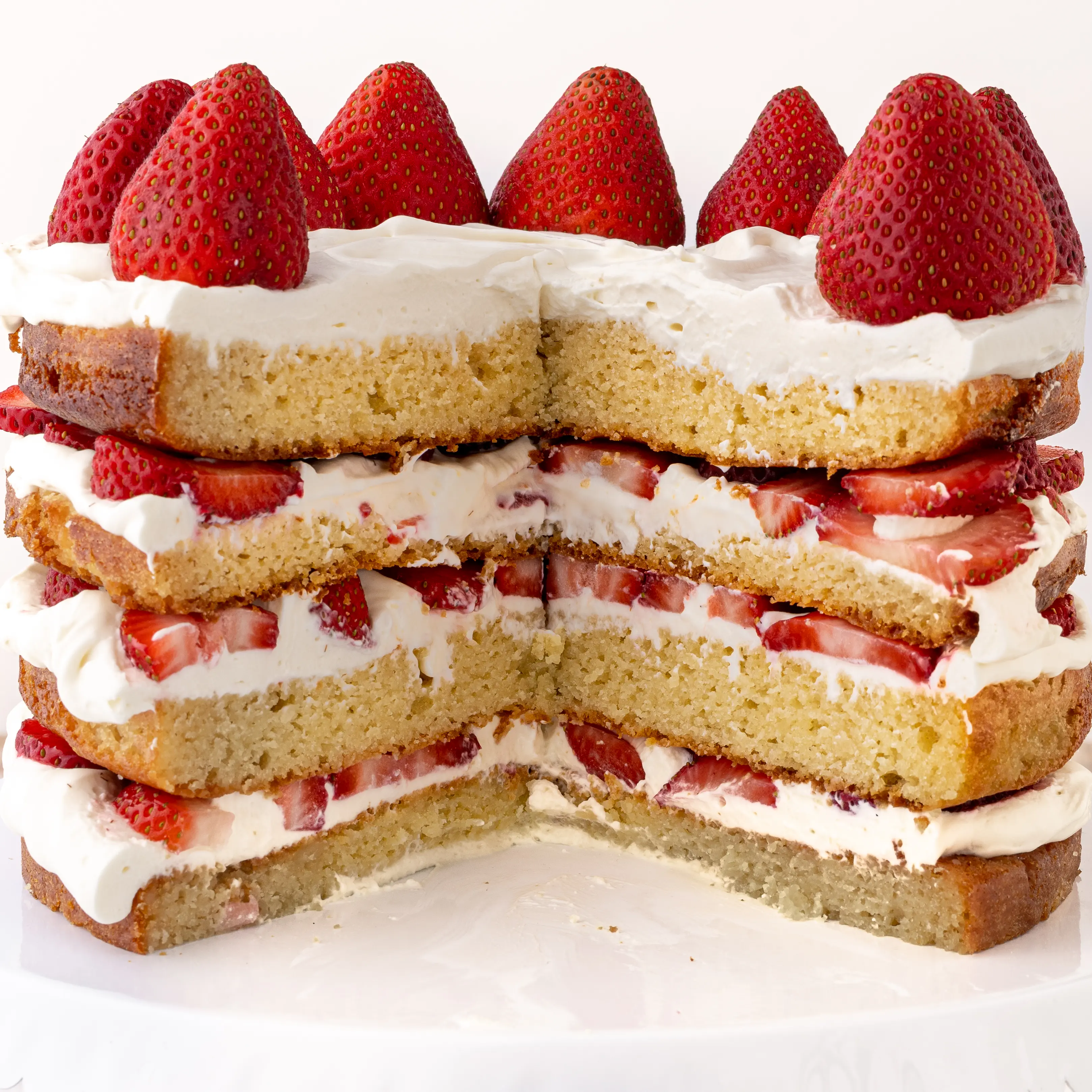 A four layer keto strawberry shortcake  sliced with fresh strawberries and whipped cream against a bright white background. 