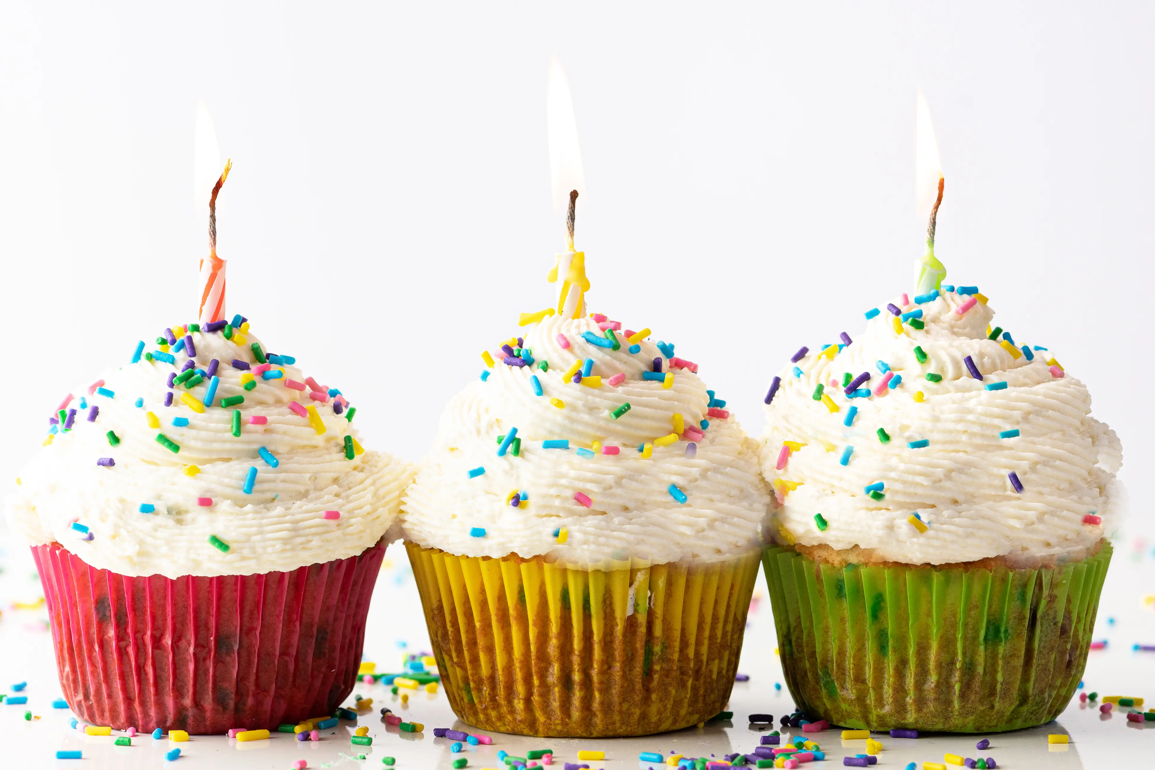 Three vanilla frosted cupcakes in colorful cupcake wrappers.  The cupcakes have cheerful sprinkles and lit birthday candles.