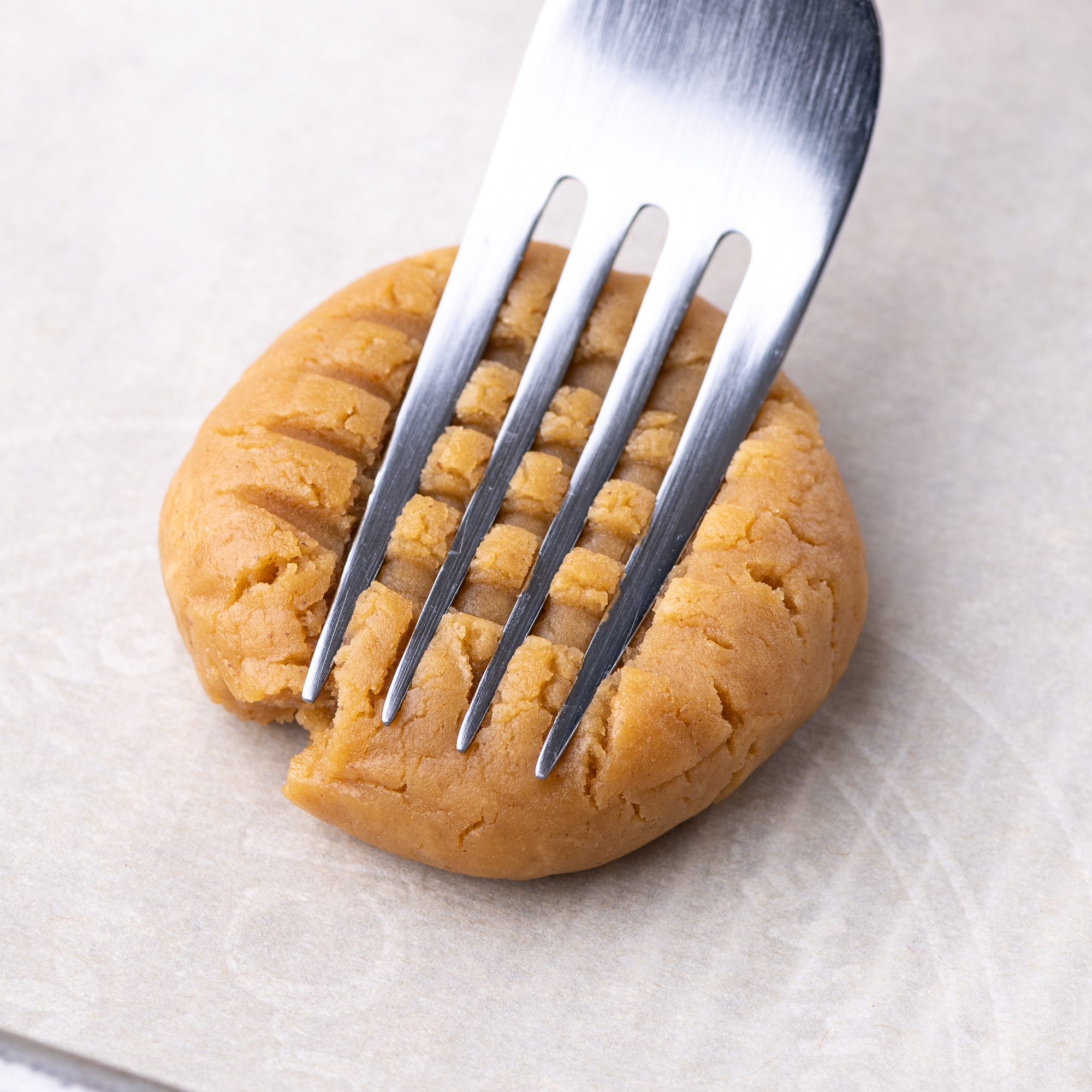 A fork being pressed into a ball of a cookie dough, creating cross hatches in the dough. 