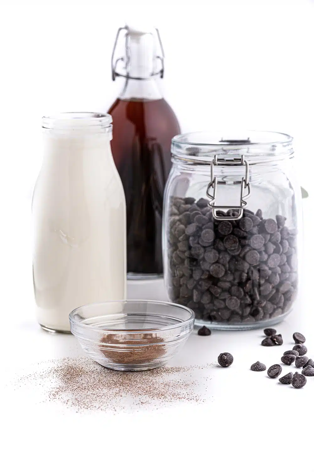 Jars filled with fresh whipping cream, chocolate chips, expresso powder and vanilla sitting on a bright white table against a bright white background. 